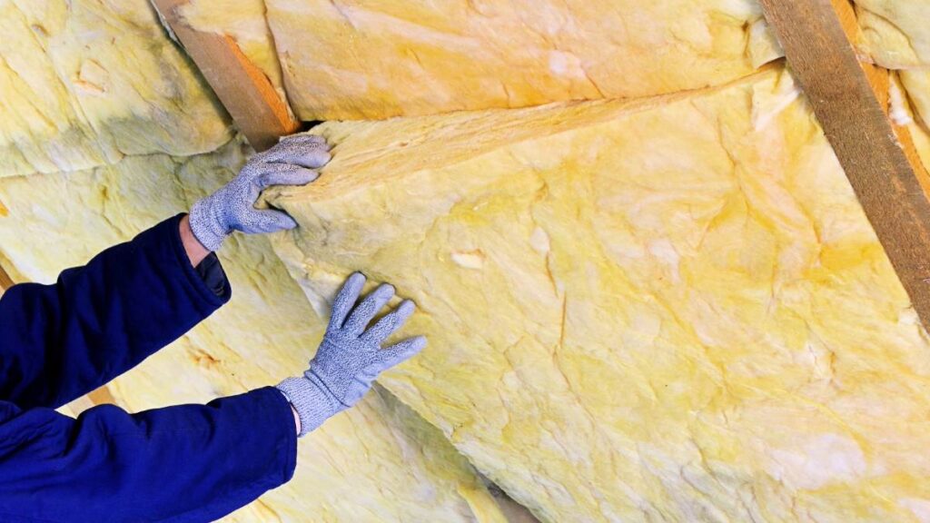 Iemand isoleert een dak met geel materiaal. Er zijn twee armen te zien die het isolatiemateriaal vasthouden. De persoon draagt grijze handschoenen en blauwe werkkleding.