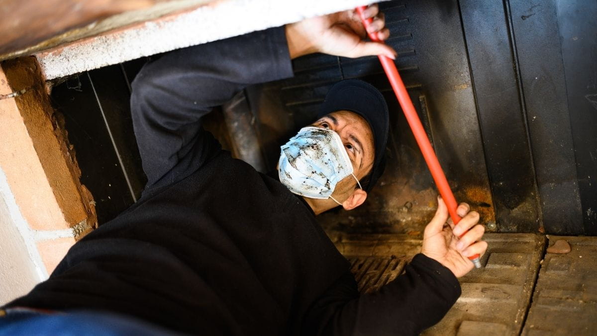 Man met stofmasker reinigt een schoorsteen van onderuit met een flexibele rode veegstok bij de open haard