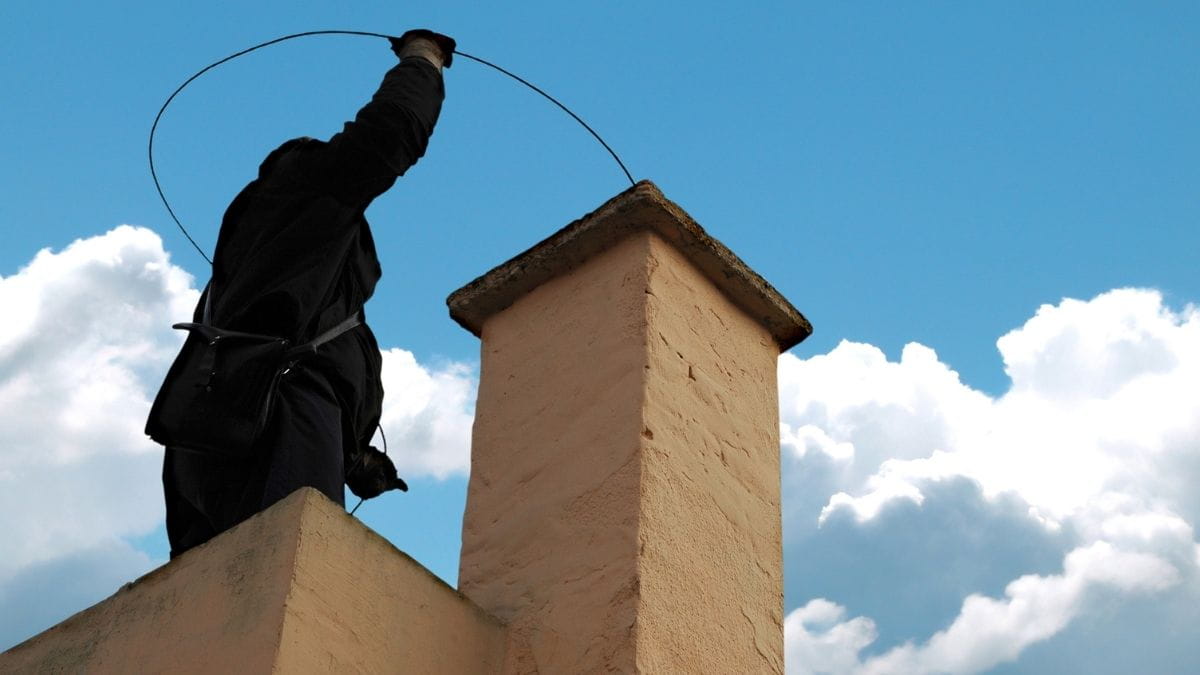 Schoorsteenveger staat op het dak en reinigt een schoorsteen met een flexibele veegstok bij een gemetselde schouw