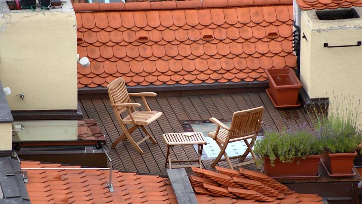 Compact houten terras met vlonderplanken en klapstoelen op een plat dak omgeven door pannendaken in de stad