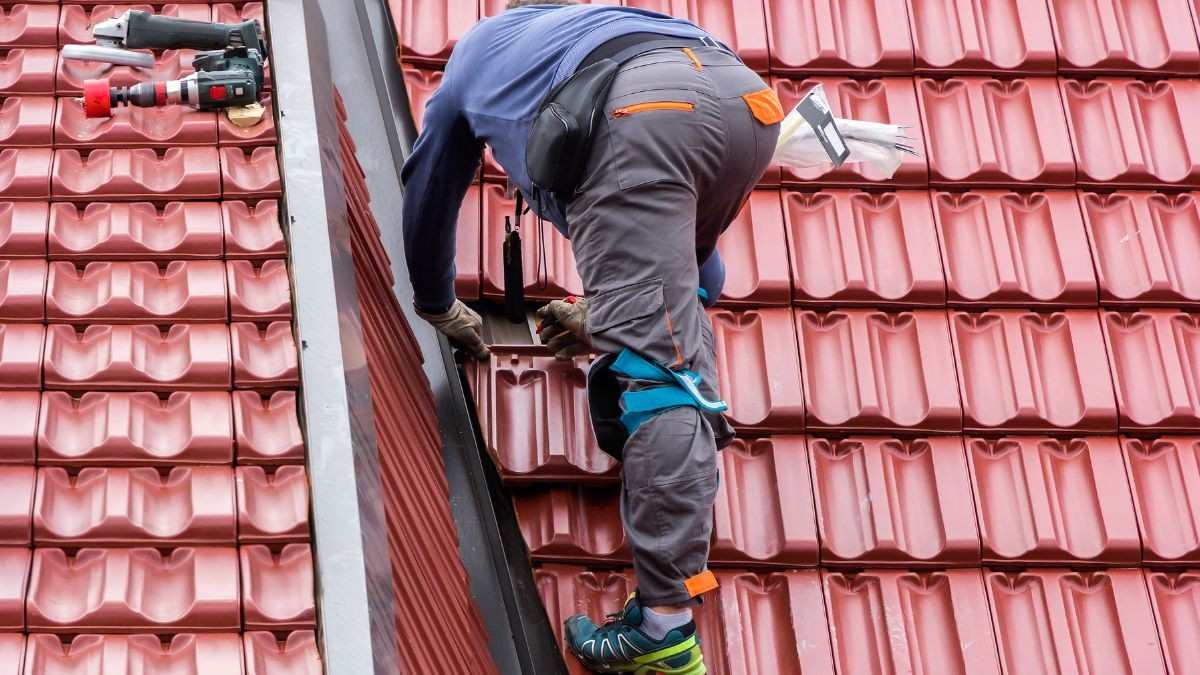 Vakman voert herstellingen uit aan rode keramische dakpannen op een hellend dak, wat de prijs van dakpannen beïnvloedt