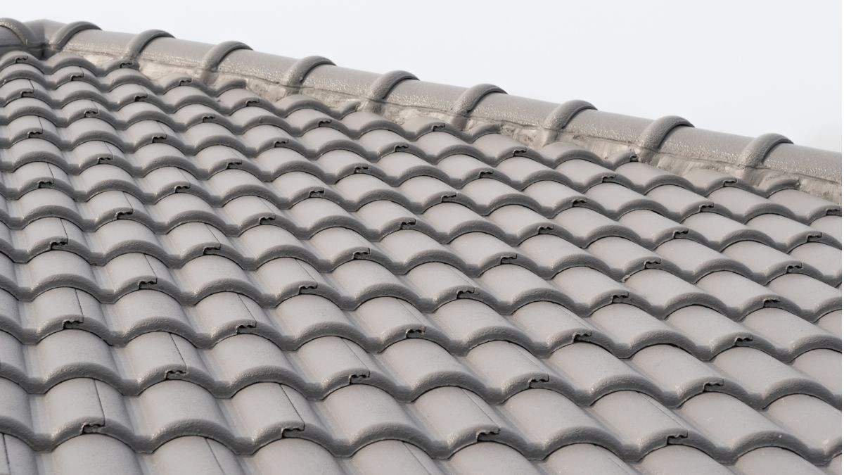 Hellend dak volledig bedekt met grijze betonnen holle dakpannen en afgewerkt met nokvorsten onder een bewolkte hemel