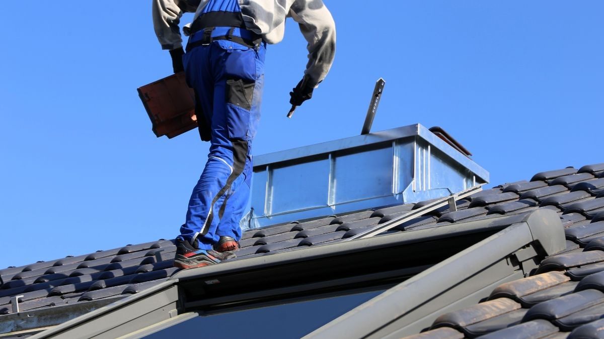 Dakwerker in blauwe werkkleding draagt een dakpan over een dak met zwarte pannen naast een geopend dakraam