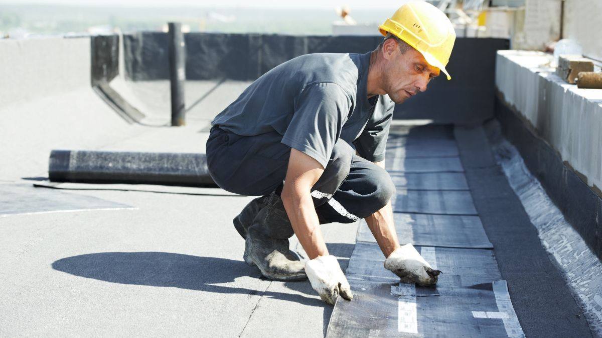 Dakwerker met veiligheidshelm legt een nieuwe bitumen afdichtingslaag op een plat dak tijdens een dakherstelling