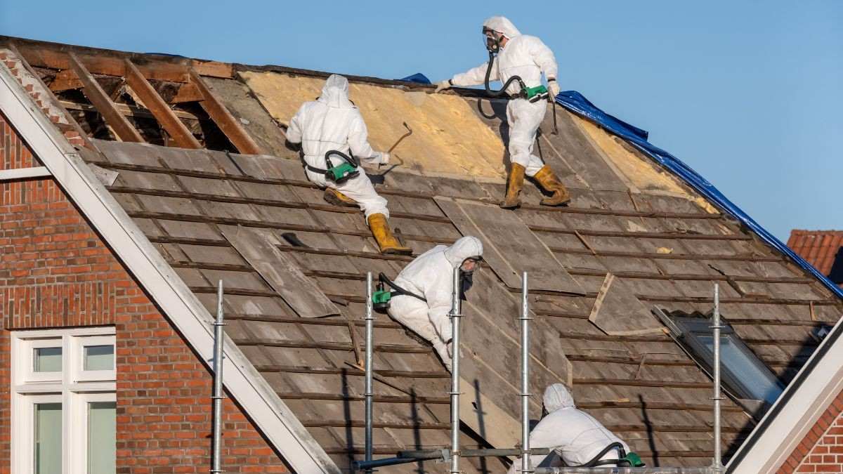 Asbestverwijderaars in beschermende pakken halen asbesthoudende dakplaten van een woning met een stelling ernaast