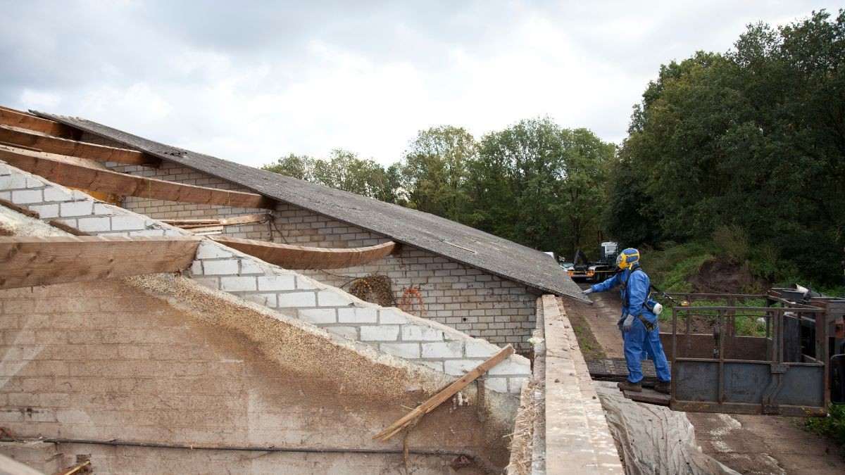 Asbestverwijderaar in beschermend pak bij een stalgebouw waar het asbestcementen dak deels al verwijderd is