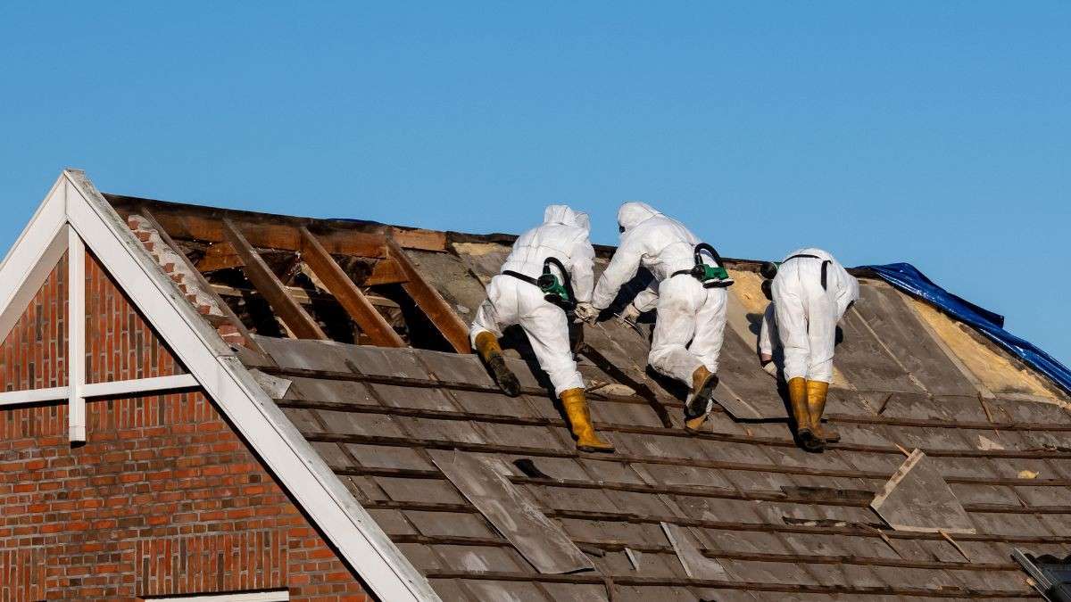 Asbestverwijderaars met ademhalingsbescherming op een steil leien dak, de hoogte bepaalt mee de asbest dak verwijderen prijs