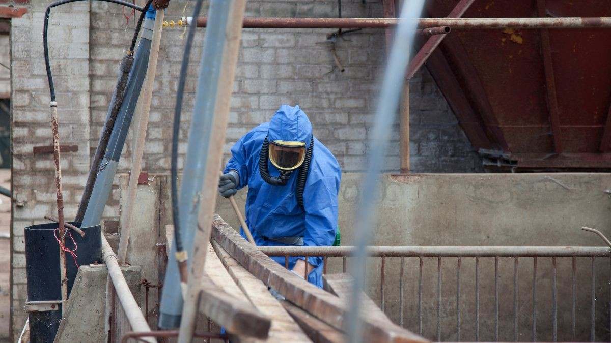Asbestverwijderaar met volgelaatsmasker en beschermingspak aan het werk in een stal van een landbouwbedrijf