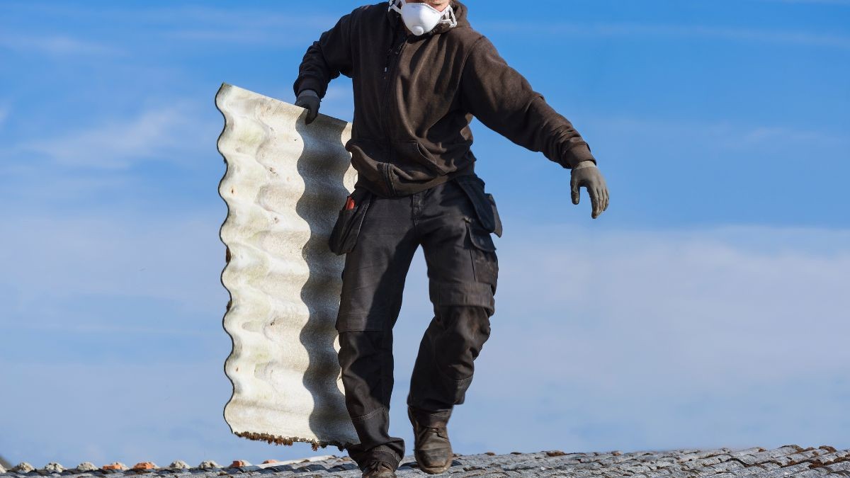 Asbestverwijderaar met stofmasker en handschoenen draagt een verweerde golfplaat in asbestcement over een oud pannendak