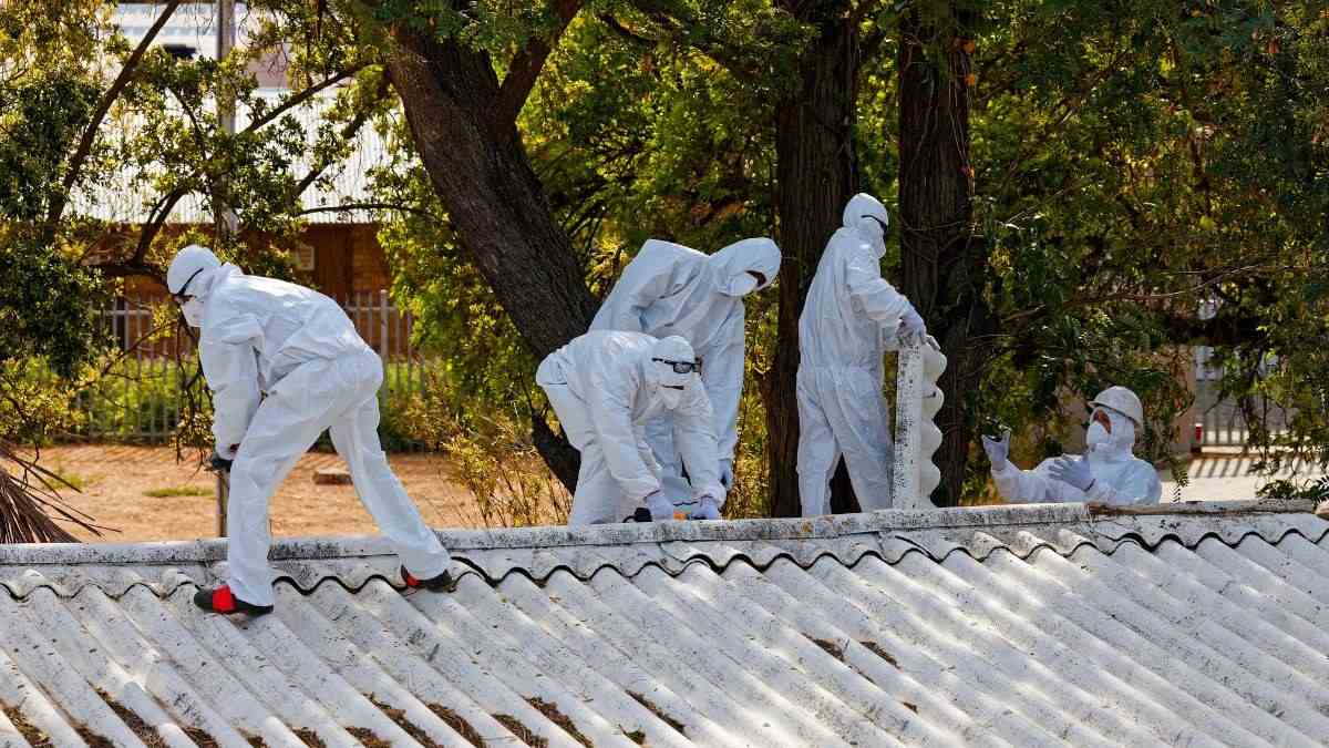 Asbestverwijderaars in witte beschermpakken halen golfplaten in asbestcement van een dak tussen de bomen, zonder ze te breken