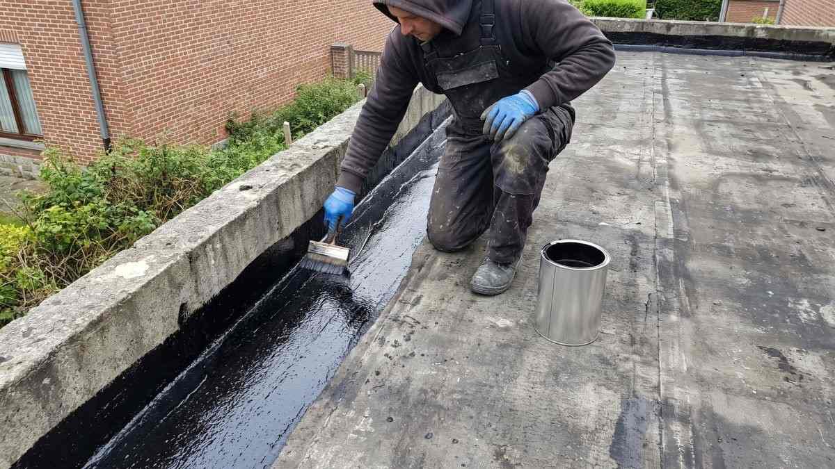 Dak coaten op een plat dak, waarbij een dakwerker zwarte bitumineuze coating met een borstel langs de dakrand aanbrengt