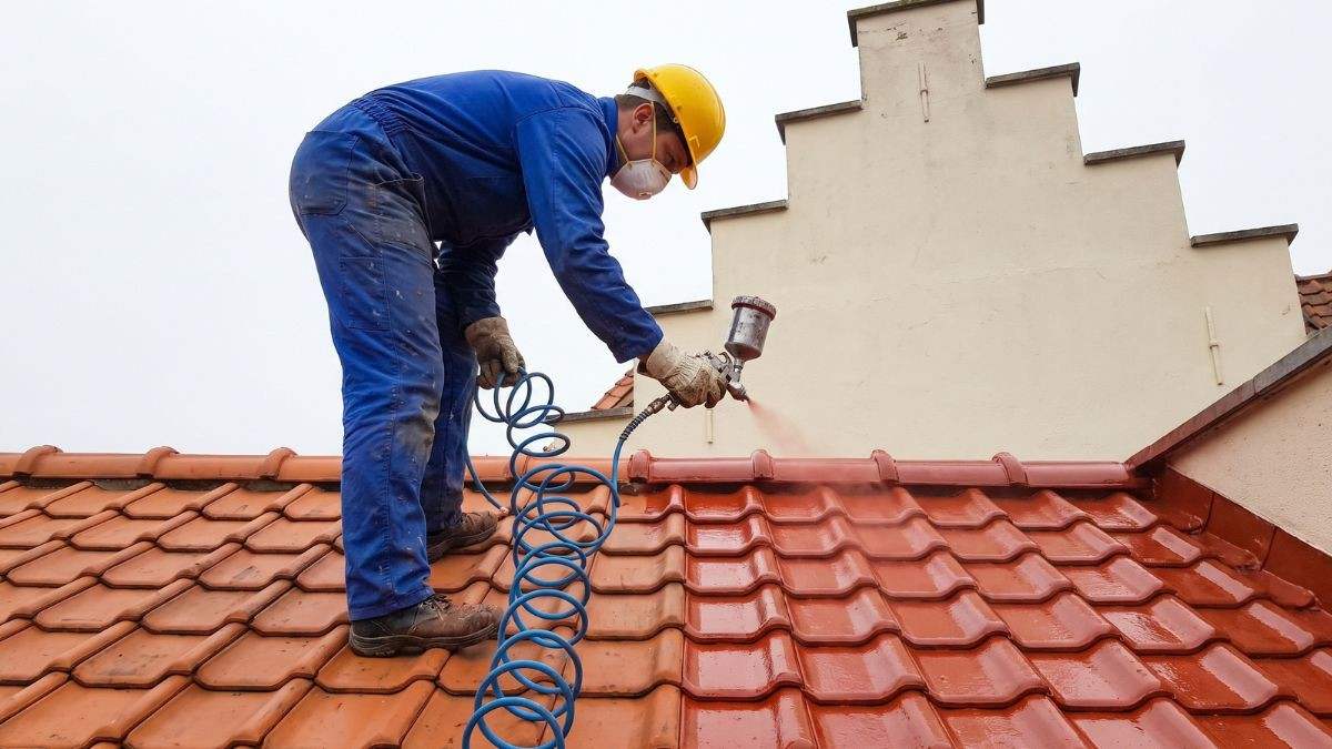 Dakwerker met helm en mondmasker die een rode coating met een spuitpistool op de kleidakpannen van een hellend dak spuit