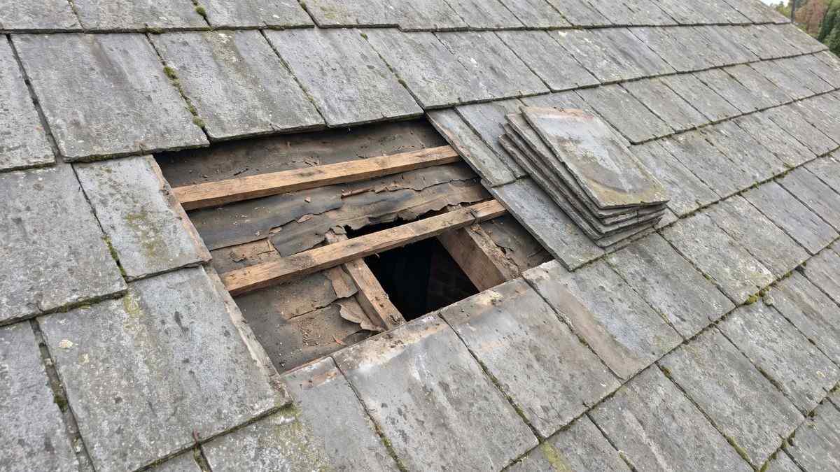 Losgemaakte kunstleien op een stapel naast een open dakvlak met zichtbare panlatten tijdens het vervangen van een oud dak