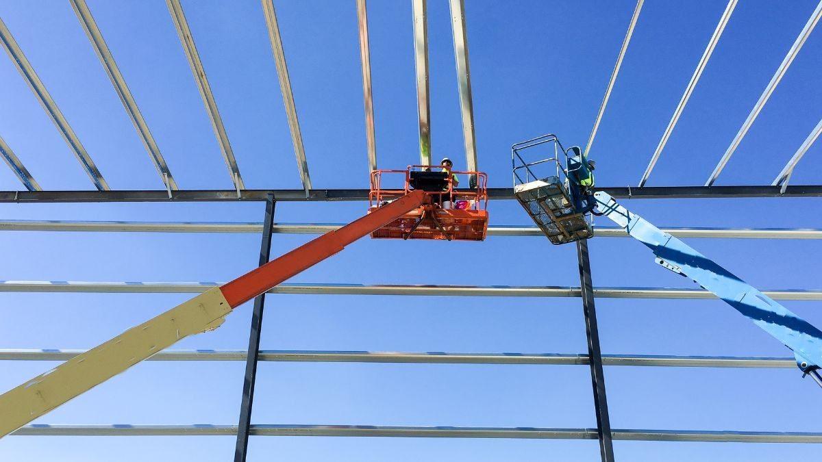 Dakwerkers op hoogwerkers monteren stalen gordingen op het draagframe van een industriële loods tegen een blauwe lucht